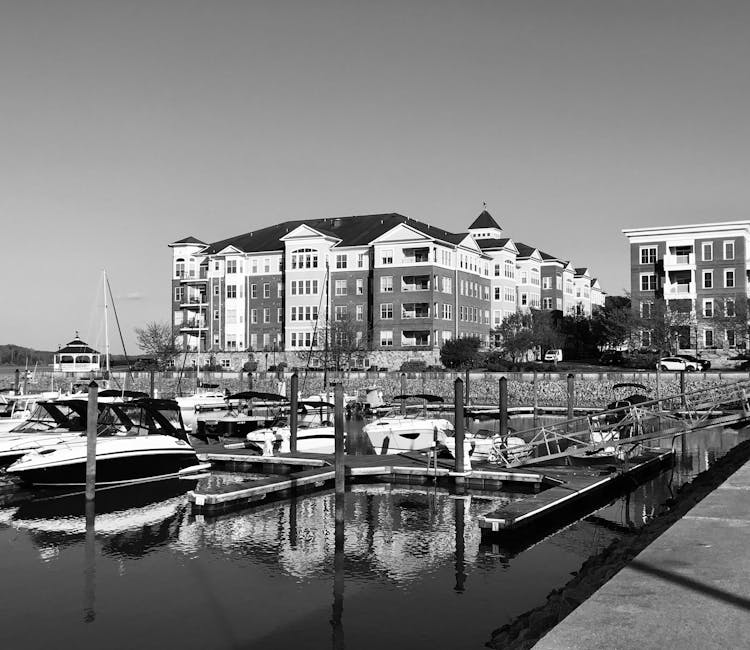 A marina scene featuring several small boats docked along wooden piers extending into calm water, with a paved walkway on the right side. In the background, there is a large, multi-story residential b