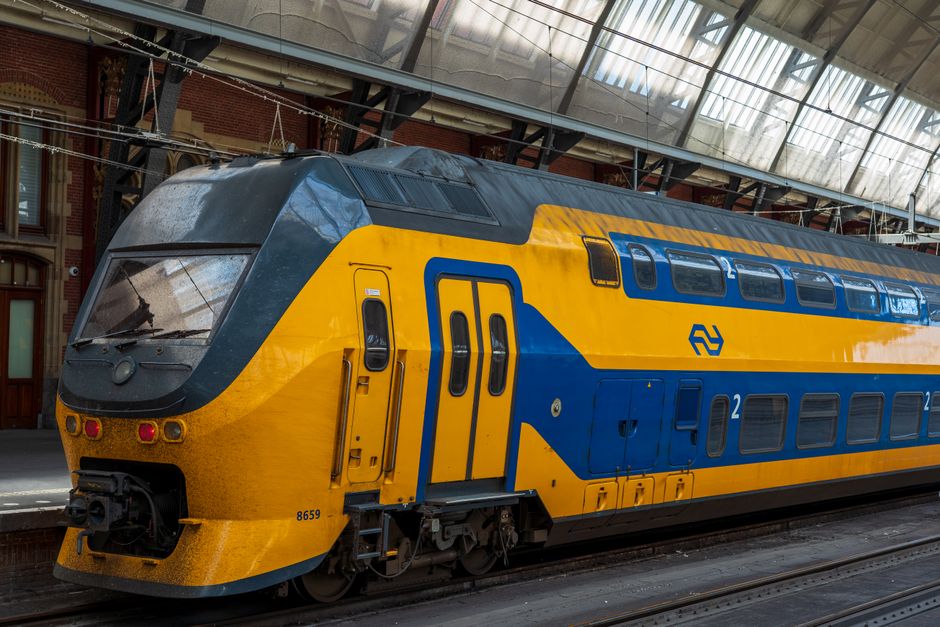 A modern double-decker train painted in bright yellow and blue, parked at Belmont Station's platform under a glass canopy. The train's sleek, aerodynamic front features large tinted windows and multip