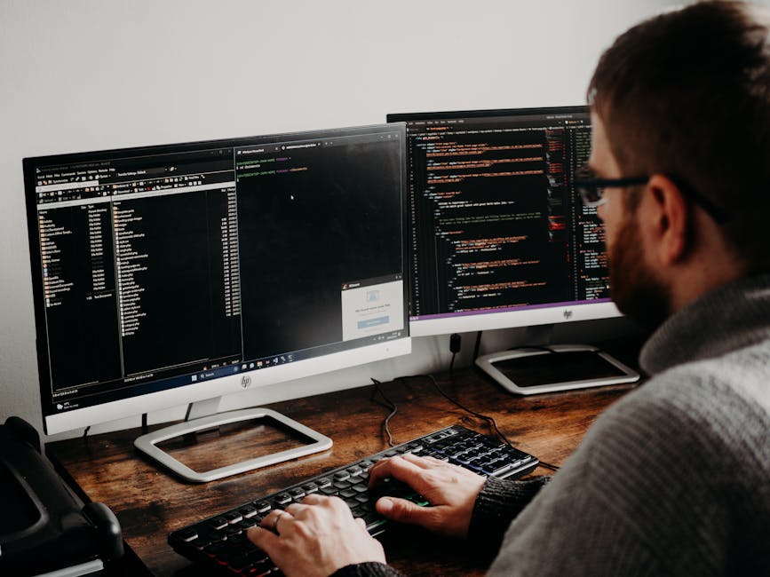 A person with a beard and glasses sits at a wooden desk working on a computer with dual monitors. The left monitor displays a file explorer and a small pop-up window, while the right monitor shows lin